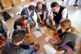 Eine Gruppe von Personen sitzt in einem Kreis auf dem Boden. Sie beschäftigen sich mit einer kreativen Aktivität, bei der sie Papier falten und Origami-Formen erstellen. Auf dem Boden liegen Zeichnungen und Anleitungen, die als Orientierung dienen. Die Teilnehmer wirken konzentriert und arbeiten gemeinsam an ihren Projekten. Stühle und weitere Materialien sind im Hintergrund sichtbar.