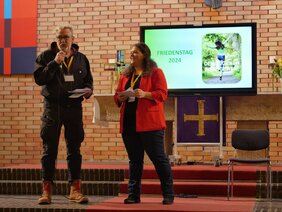 Das Bild zeigt zwei Personen auf einer Bühne in einem Kirchenraum. Eine Person, links im Bild, hält ein Mikrofon und spricht, während die andere Person, rechts, ein Blatt Papier in den Händen hält und lächelt. Im Hintergrund ist ein Bildschirm zu sehen, auf dem „Friedenstag 2024“ und das Bild einer Person in Bewegung mit dem Schriftzug „Den eigenen Weg gehen“ angezeigt werden. Die Szene wirkt wie eine Präsentation oder ein Vortrag im Rahmen einer Veranstaltung.